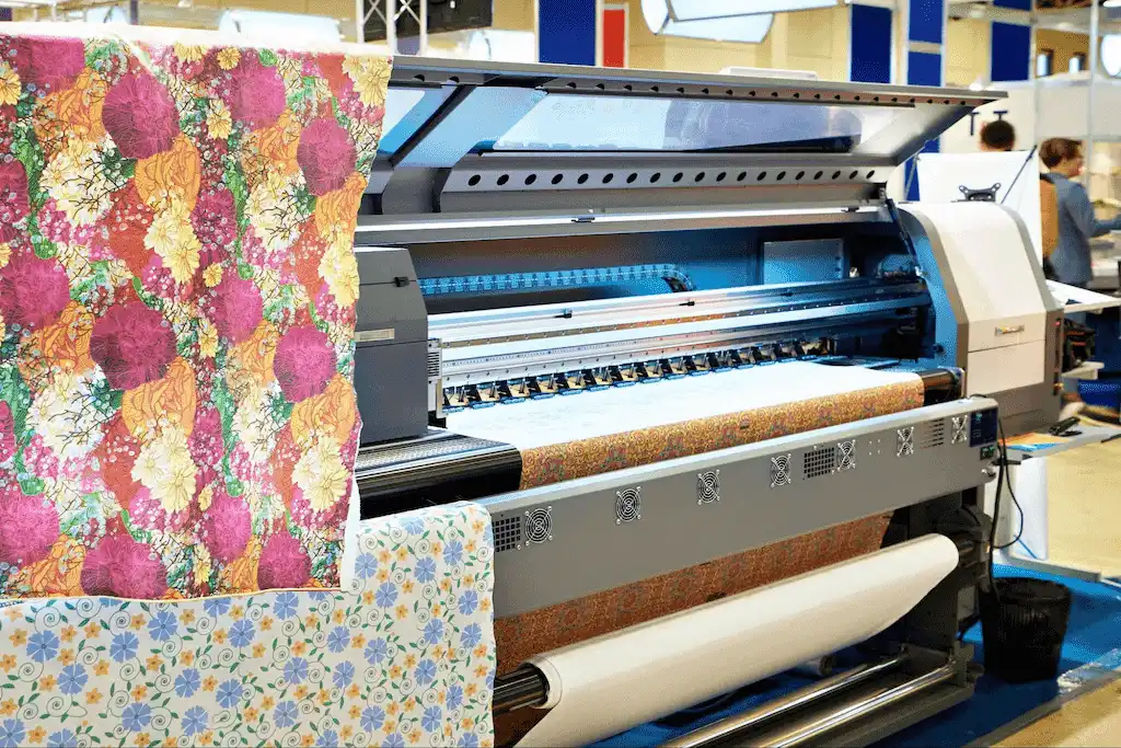 Uma grande impressora de sublimação em ação, imprimindo um design floral colorido em tecido. A imagem mostra o processo de impressão, relacionado à sublimação e ressublimação.