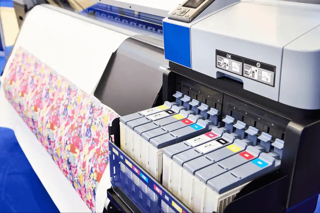 Uma impressora de sublimação com a tampa aberta, revelando o compartimento de tinta. O rolo de tecido parcialmente impresso também é visível, destacando a tecnologia por trás da sublimação e ressublimação.
